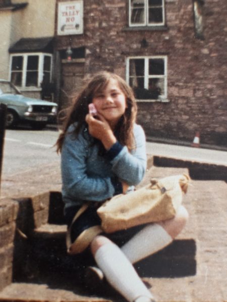 Lou, as a child, smiling at the camera sat in the square in Hatherleigh