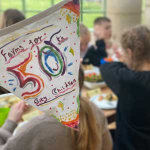 A banner celebrating Farms for City Children's 50th birthday in the foreground of a group of children eating at a table at Lower Treginnis farm.