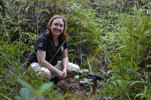 Sue smiling at the camera, kneeling with hands in front in a lush vegetation