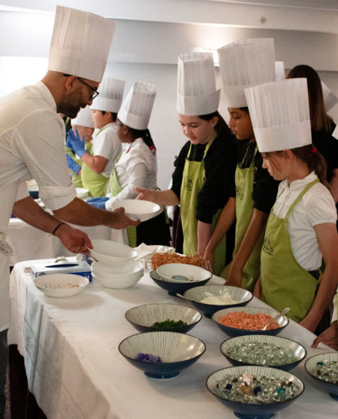 A group of young people in white chefs hat and aprons with a chef from the Savoy showing them how to make savoury canapes.