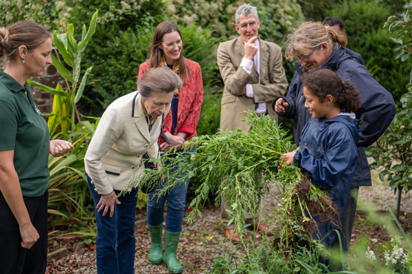 HRH The Princess Royal with a child holding a large handful of green plants in Nethercott's walled garden.