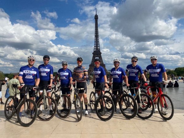 a row of cyclists in branded jerseys and bike helmets next to their bikes in a row with the Eiffel Tower in the background and fluffy cloud skies