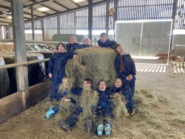 a group of children in waterproofs in a large modern barn, sat around a hay bale smiling at the camera
