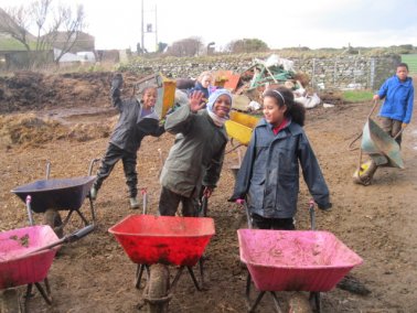 two girls in dark waterproofs and red wheelbarrows smiling at the camera in a farm yard
