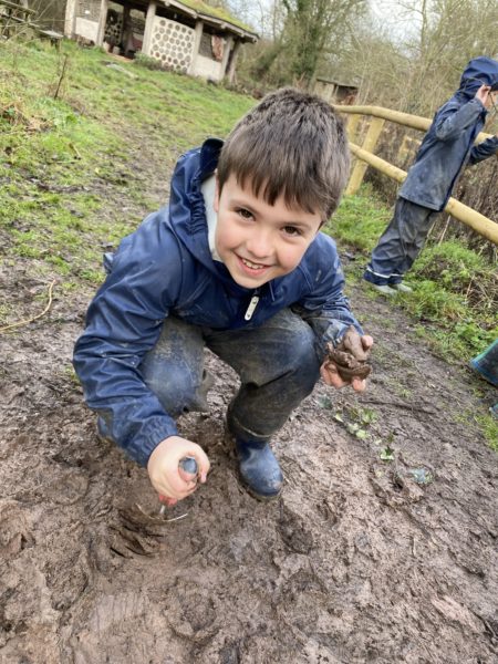A boy, in naby waterproofs crouched in the mud, smiling at the camera