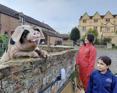 Two children in overalls looking at a pig, with Wick Court house, Farms for City Children, in the background.