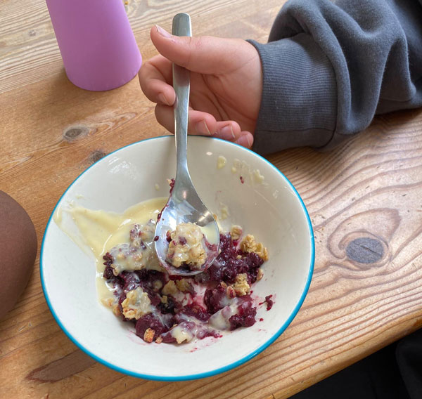 A child's hand putting a spoon into a half eaten bowl of apple and blackberry crumble.