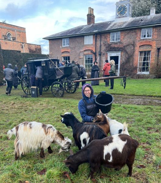 An image of sally kneeling with four goats, in a field, in front of a horse dawn carriage ad country house with a camera crew and actors