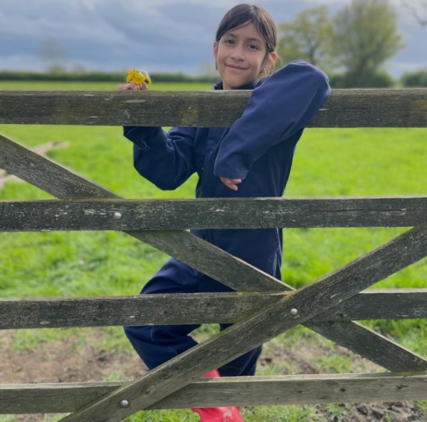 Child in farms for city children blue overalls, behind a farm gate smiling at the camera, with a posey of wild flowers in her hand