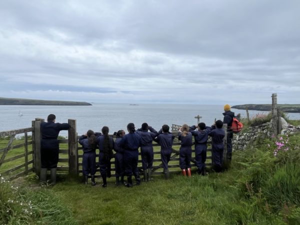children in the distance in blue waterproofs, back to the camera, leaning on a fence looking out to sea
